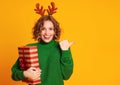 Cheerful surprised woman with deer antlers holds a Christmas gift and laughing a colored yellow background