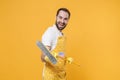 Cheerful funny young man househusband in apron rubber gloves hold in hands broom while doing housework isolated on