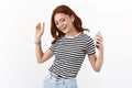 Cheerful ginger girl in striped t-shirt enjoying awesome sound quality, bought new wireless earphones, hold smartphone Royalty Free Stock Photo