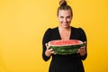 Cheerful girl in black dress joyfully looking in camera while holding half of big watermelon in hands over yellow