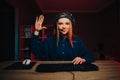 Cheerful girl gamer in a headset on her head and in casual clothes plays video games at home on the computer, shows a greeting Royalty Free Stock Photo