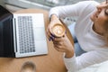 Cheerful girl holding a cup of fragrant cappuccino with froth while working at laptop in a cafe. Royalty Free Stock Photo