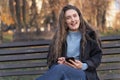 Cheerful girl with long brown hair sits in park on bench and uses her phone. Young woman with smartphone outside Royalty Free Stock Photo