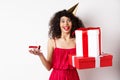Cheerful girl in party hat and red dress, celebrating birthday, holding surprise gift and b-day cake, enjoying holiday Royalty Free Stock Photo