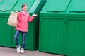 Cheerful girl in a pink jacket, carries a paper bag in a green garbage container to throw it away Royalty Free Stock Photo