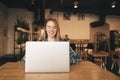 Cheerful girl sits in a cozy cafe with a laptop, looks into the camera and smiles. Attractive girl student enjoys a laptop, Royalty Free Stock Photo