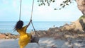 Cheerful girl swinging on a swing on a tropical beach with an ocean view