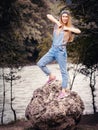 A cheerful girl taps her fists on her chest standing on a round stone against the background of the river on a summer