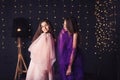 Cheerful girlfriends. Two long-haired girls in pink and purple dresses in studio on dark background with bokeh. Copy space. Royalty Free Stock Photo