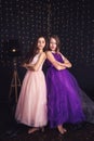 Cheerful girlfriends. Two long-haired girls in pink and purple dresses in studio on dark background with bokeh. Copy space.