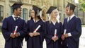 Cheerful graduate students in academic regalia talking to each other, success