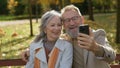 Cheerful gray-haired wife husband using smartphone online communication on bench in autumn park nature old grandparents Royalty Free Stock Photo