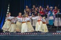 Cheerful group of children wearing traditional Polish costumes dance on stage.