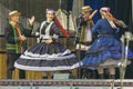 Cheerful group of children wearing traditional Polish costumes dance on stage.