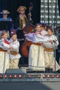 Cheerful group of children wearing traditional Polish costumes dance on stage.