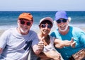 Cheerful group of three friends sitting on the beach, retired adult people in outdoor vacation at sea smiling enjoying sun and Royalty Free Stock Photo