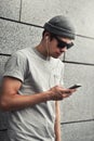 Cheerful guy dressed in gray t-shirt, sunglasses and hat at the street, listening to music with earphones, holding mobile phone. Royalty Free Stock Photo