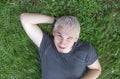 A cheerful guy in a gray T-shirt is lying on a green lawn with one eye squinted