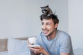 Cheerful handsome man with cute raccoon on head holding coffee cup while sitting on bedding. Royalty Free Stock Photo