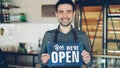 Cheerful handsome man small coffee-house owner is holding We Are Open sign while standing inside coffee shop. Opening Royalty Free Stock Photo