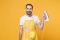 Cheerful handsome young man househusband in apron hold in hands iron while doing housework isolated on yellow wall