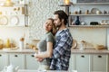 Cheerful happy caucasian millennial hasband with stubble hugs blond wife, enjoy tender moment in light kitchen