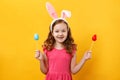 Cheerful happy child in the ears of a bunny holds two Easter eggs on a stick. Portrait of a little girl on a yellow background Royalty Free Stock Photo