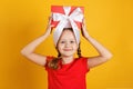 Cheerful happy little girl holds a box with a gift on her head. Child in a red T-shirt on a yellow background Royalty Free Stock Photo