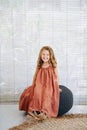 Cheerful happy little girl in long dress sitting on a high black wicker pillow Royalty Free Stock Photo