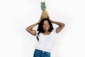 Cheerful happy pretty young african woman in white t-shirt and jeans, holding a pineapple on her head, looking at camera Royalty Free Stock Photo