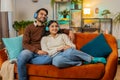 Cheerful happy young Indian couple in wireless headphones dancing on couch at home listening music Royalty Free Stock Photo