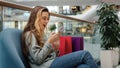 Cheerful happy young girl shopper shopaholic woman sitting in shopping mall with packages looking at phone wins rejoices