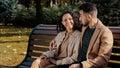 Cheerful hispanic couple cutely communicate sitting on bench in autumn city park young bearded guy hugs attractive curly Royalty Free Stock Photo