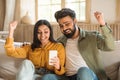 Cheerful indian couple using smartphone and shaking fists, reading great news, celebrating big luck at home Royalty Free Stock Photo