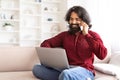 Cheerful indian guy using laptop, talking on phone at home Royalty Free Stock Photo