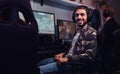A young cheerful Indian guy wearing a military shirt sitting on a gamer chair and looking at a camera in a gaming club