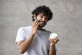 Cheerful indian man applying cream on face, holding moisturizer jar in hand, smiling to camera, standing in bathroom