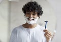 Cheerful indian man with face covered with shaving foam holding safety razor while looking at mirror in bathroom Royalty Free Stock Photo