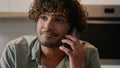 Cheerful Indian young man with curly hair talking phone in home kitchen. Happy friendly Hispanic guy talk with friends Royalty Free Stock Photo