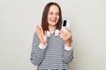 Cheerful joyful brown haired adult woman wearing striped shirt standing isolated over gray background holding smart phone having