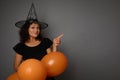 Cheerful joyful happy Hispanic woman in wizard hat, dressed in black, holds orange inflated air balloons and points on a copy Royalty Free Stock Photo