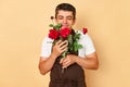 Cheerful joyful male florist wearing brown apron standing isolated over beige background at flowers shop smelling beautiful red Royalty Free Stock Photo