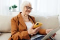 a cheerful, joyful old lady in a brown pantsuit is sitting on a cozy sofa holding a smartphone in her hand and a laptop Royalty Free Stock Photo