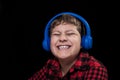 Cheerful smiling young boy in a plaid shirt listening to music in big blue wireless over-ear headphones on isolated black Royalty Free Stock Photo