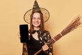 Cheerful joyful young woman wizard wearing witch costume holding in hand broom  over beige background standing showing Royalty Free Stock Photo