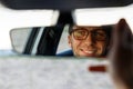 Cheerful joyfull man wearing glasses and adjusting mirror while sitting in his car, looking in reflection. soft focus