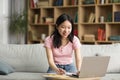 Cheerful korean lady attending webinar from home, sitting on sofa, using laptop and taking notes, copy space