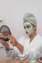 cheerful lady enjoys a pampering day with green clay mask, smiling in the mirror's reflection.