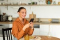 Cheerful lady using smartphone while sitting at kitchen table and having lunch, texting on mobile phone, free space Royalty Free Stock Photo