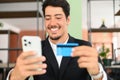 Cheerful latin man makes purchase online. Hispanic guy in business wear holds phone and a credit card Royalty Free Stock Photo